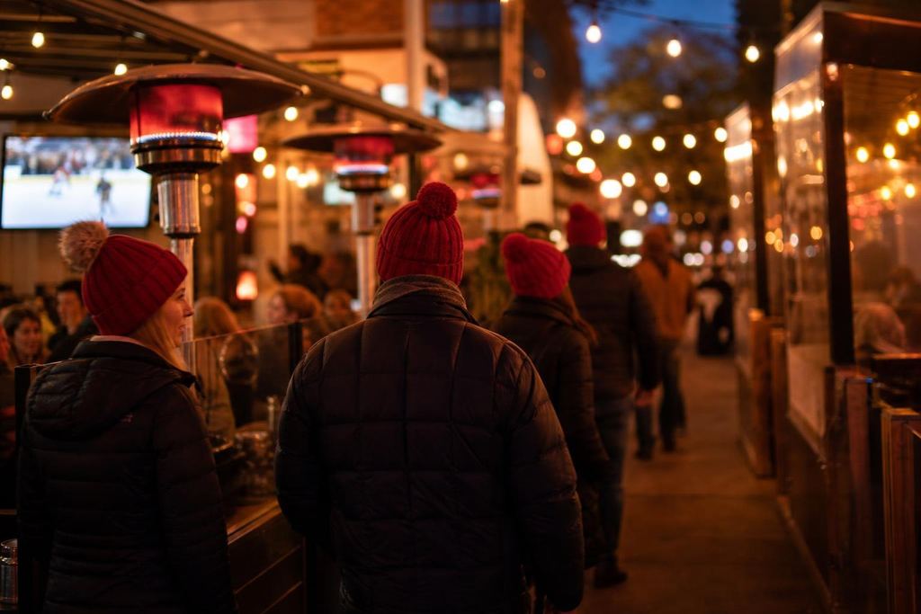An avenue of bars at night with heated patios, string lights and fans in red toques (AI-generated image)