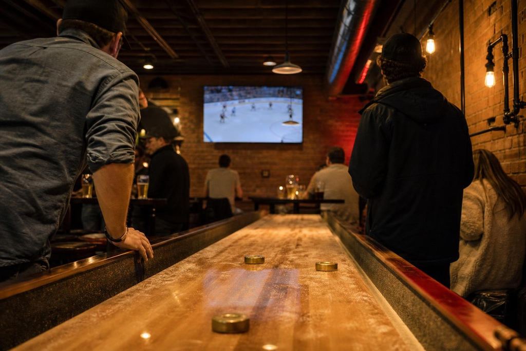 A long wooden shuffleboard table in a brick-walled basement bar under warm pendant lights (AI-generated image)